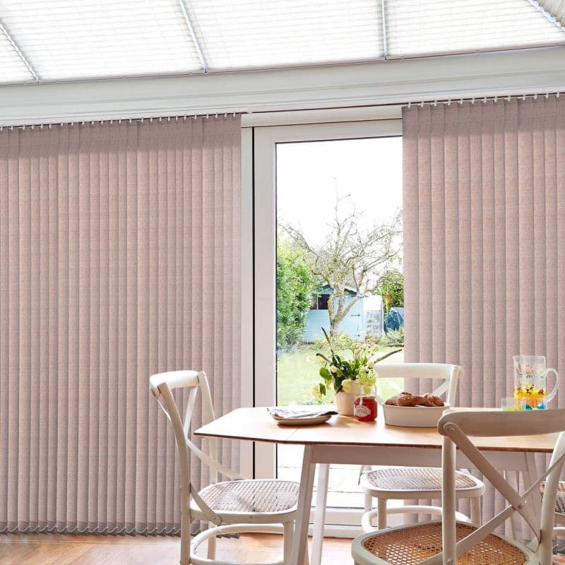 Beige vertical fabric blinds covering a sliding glass door, mostly closed with a central gap; filtering daylight into a small dining area with wooden table, chairs, vase, and backyard view.