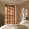 Wooden plantation shutters covering a tall window—four full-height panels with adjustable slats—partially closed, casting striped sunlight across a softly lit bedroom with bed, nightstand, patterned wallpaper and dresser.