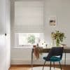 Patterned fabric Roman blind lowered over a sash window, softly diffusing daylight; a wooden desk with teal chair and vase of flowers sits beneath in a neutral, sunlit study.