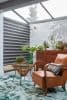 Striped dual-layer roller blind covering left glass wall, alternating dark and light horizontal bands, partially closed filtering daylight; sunroom with glass ceiling, leather armchair, rattan table, plants and wooden chest.