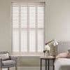 White four-panel plantation shutters covering tall window, horizontal louvers closed but slightly angled, filtering soft daylight; bedroom corner with upholstered bed, bedside table, lamp, vase of flowers and armchair.