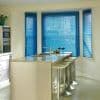 Blue horizontal Venetian blinds covering a three-panel bay window, partially closed and filtering daylight; modern kitchen with marble-topped island, stainless-steel sink and three metal bar stools, bright neutral walls.
