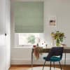 Sage-green fabric Roman blind, partially lowered with soft folds, filtering daylight through a white sash window; scene shows a wooden desk with teal chair, vase of flowers, mirror, pale walls.