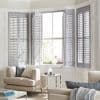 White wooden plantation shutters on a bay window; outer panels closed, center panels partially open, slats filtering bright daylight into a sunlit living room with beige sofas and side table.