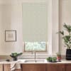 Patterned roller blind covers most of a kitchen window above a stainless sink, set amid a wooden-counter kitchen with faucet, bowl of greens, a towel, framed sketch, and potted plants.