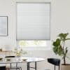 White pleated blind lowered over a recessed window, filtering soft daylight; dining table with glassware and vase, black chairs and a fiddle‑leaf fig plant in a bright minimalist room.