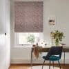 Patterned Roman blind (red leafy print) lowered over a sash window, filtering daylight; wooden desk beneath with mirror, teal chair and vase of flowers in a bright, neutral-toned home office.