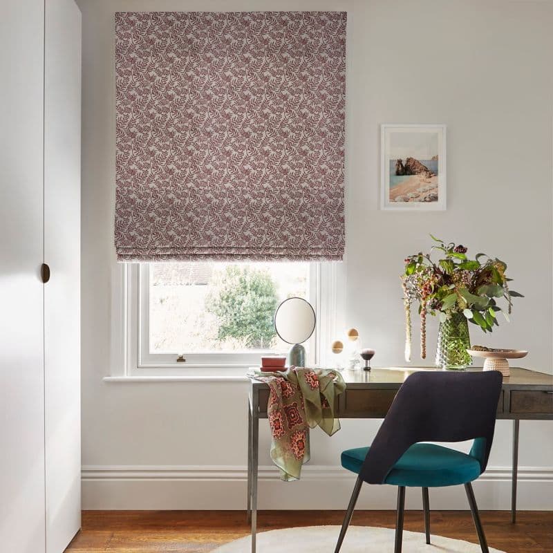 Patterned Roman blind (red leafy print) lowered over a sash window, filtering daylight; wooden desk beneath with mirror, teal chair and vase of flowers in a bright, neutral-toned home office.