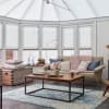 White pleated cellular blinds covering bay windows and glazed skylight, lowered to diffuse daylight; conservatory seating area with beige sofa, chunky pink throw, wooden coffee table and blue-pattern rug beneath.