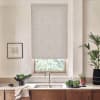 Beige patterned roller blind covers most of a kitchen window above a sink with stainless faucet; wooden cabinets and countertop display bowls of leafy greens, potted plants, and framed sketch.