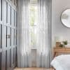 Sheer patterned floor-length curtains, slightly parted over a sash window, filtering soft daylight; bedroom with dark paneled wardrobe to the left, wooden bedside table, round mirror and greenery outside.