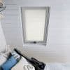 White pleated skylight blind, fully lowered with horizontal folds, filtering soft daylight; set in a white-painted attic with slatted ceiling above a bed with blue, patterned, and guitar-shaped pillows.