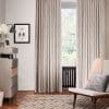 Beige patterned pleated curtains, hung floor-to-ceiling and drawn closed at sides leaving central pane exposed, filtering soft daylight; living-room setting with radiator beneath window, armchair, sideboard and patterned rug.