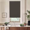 A dark roller blind covers most of a kitchen window, blocking light above a sink; countertop holds bowls of leafy greens, a dish towel, framed sketch on the wall, and potted plants.