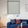 Light-gray cellular blind, horizontally pleated, fully lowered over a window, filtering daylight; in a sunlit living room with a blue sofa, assorted cushions, a floor lamp and small coffee table.