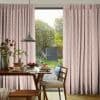 Pleated pink diamond-pattern curtains, drawn apart over a sliding glass door; filtering soft daylight into a dining room with wooden table, pendant lamp and a sunlit garden view.