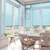 Light-blue horizontal venetian blinds covering tall floor-to-ceiling windows, largely lowered and filtering soft daylight; bright white dining sunroom with wooden table, chairs and garden view.