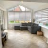 Zebra roller blinds covering triangular-topped bay and side windows, lowered and filtering daylight in a bright living room with a grey corner sofa, wood herringbone floor, overlooking a lawn.