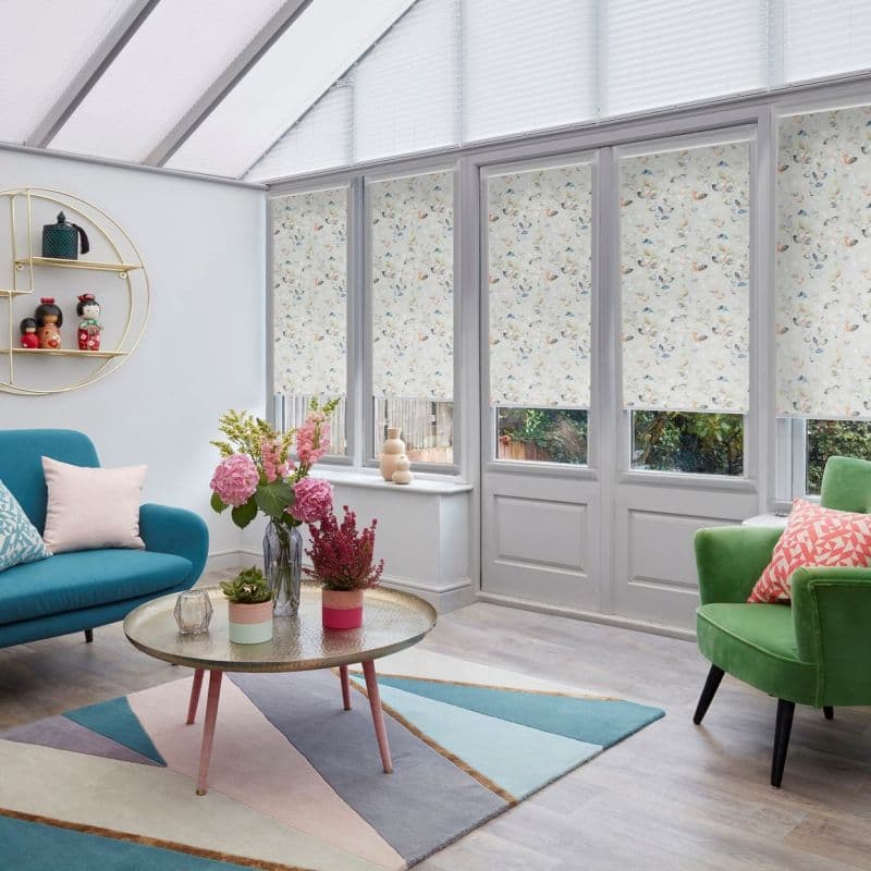 Floral roller blinds on tall windows and doors, partially lowered and filtering daylight; pleated blinds cover the sloped roof. Bright conservatory-style living room with colorful sofas, rug, and coffee table.