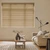 Horizontal beige Venetian blinds cover a large window, closed and gently filtering daylight; a neutral living room with a sofa, wooden coffee table, patterned rug and angled black floor lamp.