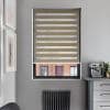Zebra roller blind, beige alternating opaque-and-sheer horizontal bands, lowered across a recessed window; filtering light, partially revealing a brick building, in a gray kitchen corner with radiator and espresso machine.