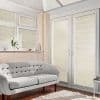 White pleated blinds on French doors and adjacent windows, fully closed and filtering sunlight, casting horizontal shadows across a tufted sofa in a bright living room with a shag rug.