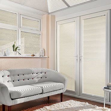 White pleated blinds on French doors and adjacent windows, fully closed and filtering sunlight, casting horizontal shadows across a tufted sofa in a bright living room with a shag rug.