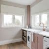 White Roman blinds, lowered and softly folded across two windows, filtering overcast daylight; laundry room countertop with pale‑pink cabinets, sink, washing machines, wooden floor and a view of bare trees.
Whirlpool