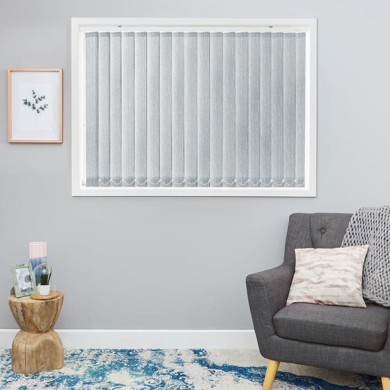 Vertical grey fabric blinds, closed and softly diffusing light; in a window — neutral living room with grey walls, botanical print, wooden stump side table, grey armchair and blue rug.