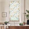 Floral-patterned roller blind covers a kitchen window, partially lowered above a stainless sink; countertop holds bowls of greens, potted plants, a towel, and a framed sketch on the adjacent wall.