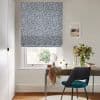 A navy-and-white geometric Roman blind, lowered and filtering soft daylight over a window; a desk beneath holds a mirror, vase of flowers, and a teal chair in a neutral office.