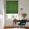 Green Roman blind — solid fabric with soft horizontal folds — is lowered, filtering daylight; a neat desk and teal chair sit beneath in a bright, minimalist home office with flowers.