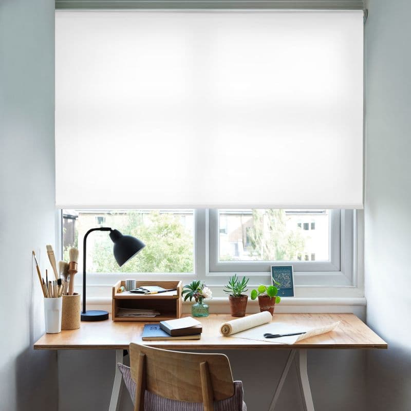 White roller blind, lowered across a double window, opaque and diffusing bright daylight; covering most of the pane above a tidy wooden desk with lamp, brushes, potted succulents, and suburban view.
"British Herbs"