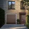 Sheer curtains in the upper-left black-framed window, partly drawn and filtering daylight; set on a brick townhouse facade with matching brown garage and front door, potted shrub and shaded driveway.