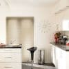 Beige horizontal Venetian blinds, fully closed, diffusing soft daylight through kitchen window; white kitchen with grey countertops, chrome trash bin, black barstool, wall clock and red flowers on counter.