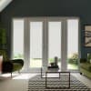 Glass patio doors and flanking tall windows covered by lowered white roller blinds, framed by green armchairs; glass coffee table on black-and-white chevron rug; sunlit garden glimpsed beneath blinds.