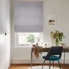 A light grey roman blind lowered over a sash window, soft-folded fabric gently filtering daylight; below, a desk with mirror, vase of flowers and teal-upholstered chair in a home office.
