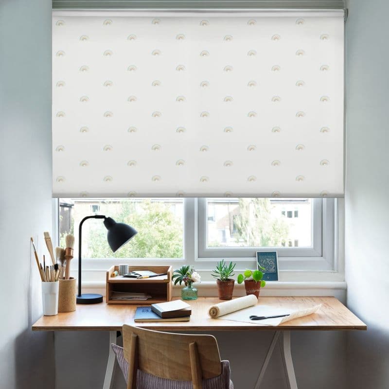 A white roller blind printed with small pastel rainbows, lowered over a window, filtering soft daylight onto a wooden desk with lamp, art supplies and plants in a bright workspace.