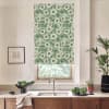 Green floral roller blind hangs partially lowered over a window above a sink and faucet in a kitchen, surrounded by wooden cabinets, potted plants, a black bowl and framed wall art.