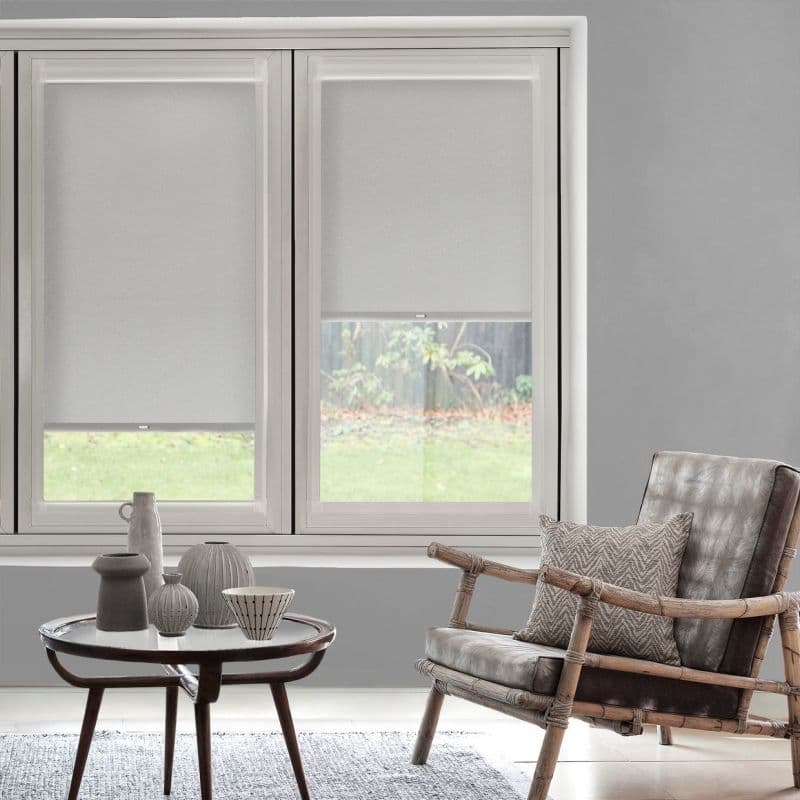 White roller blinds on twin windows, lowered to mid-height and filtering daylight; interior is a grey living room with rattan armchair, patterned cushion, round coffee table and backyard view.