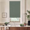 A patterned green roller blind covers a kitchen window, lowered halfway. A stainless faucet and double sink sit below on a marble countertop, flanked by plants, vegetables, and framed artwork.
