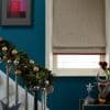 Beige Roman blind with subtle texture and red fringe, lowered over a white-framed window, softly diffusing daylight in a teal-walled hallway beside a garland-wrapped staircase and metallic holiday ornaments.