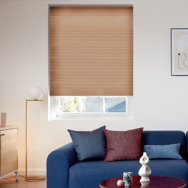 Honey‑tan pleated cellular blind, lowered and closed with fine horizontal ridges and a slight bottom gap, filtering daylight; modern living room with blue sofa, colored cushions, floor lamp and sideboard.
