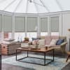 Light-gray cellular (honeycomb) blinds, lowered over conservatory windows, filtering diffuse daylight; in a bright sunroom with a beige sofa, wooden coffee table, blue-pattern rug and decorative cushions.