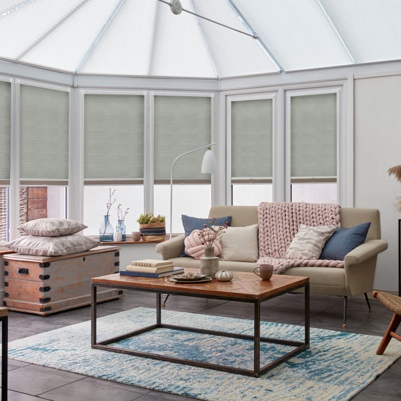 Light-gray cellular (honeycomb) blinds, lowered over conservatory windows, filtering diffuse daylight; in a bright sunroom with a beige sofa, wooden coffee table, blue-pattern rug and decorative cushions.