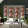 Floral-patterned roller blinds cover four tall glass doors, filtering daylight in a modern living room furnished with green armchairs, a glass coffee table and a chevron rug.