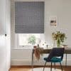 Navy-and-white geometric Roman blind, lowered over most of the window, filtering daylight; a wooden desk beneath in a bright workspace with a teal chair, vase of flowers, and framed print.