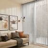 Sheer vertical blinds, ceiling-to-floor pale-gray slats covering a sliding glass door, filtering soft daylight and casting striped shadows; neutral living room with beige sofa, framed art and wall lamp.