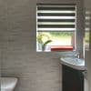 Dual-layer zebra roller blind, horizontal opaque-and-sheer stripes, partially lowered, filtering daylight; frosted sill holds potted orchid and folded orange towel in tiled bathroom with corner sink, toilet.