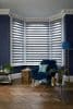 Zebra roller blinds on a three-panel bay window, alternating sheer and opaque stripes, partially closed and diffusing daylight; navy living-room corner with velvet armchair, gold accents, and parquet floor.